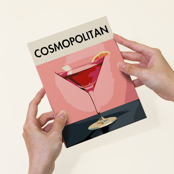 Sip Society paint by numbers kit by Artspark featuring the finished Cosmopolitan canvas, held by hands. Stylized cocktail illustration with a pink background and a lemon twist garnish.