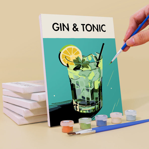 Gin & Tonic canvas from the Sip Society paint by numbers set by Artspark. Close-up of the painting process with a brush applying green paint to a stylized glass of gin and tonic with lemon slice and mint leaves on a teal background. Includes paint pots, brushes, and stacked canvases in the background.