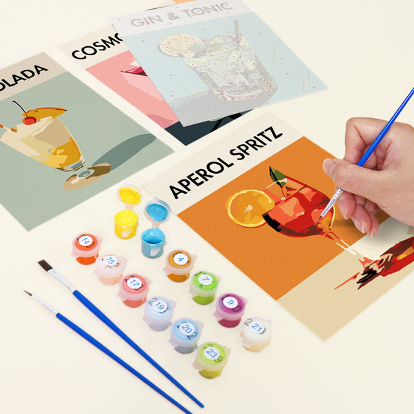 Sip Society paint by numbers kit by Artspark showing the painting process with Aperol Spritz canvas, unpainted Gin & Tonic canvas, paint pots, and brushes. Multiple cocktail designs in progress.
