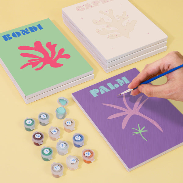 Coastal Dreams Paint by Numbers Kit by Artspark showing the painting process of the Palm canvas with paint pots, brush, and stacked canvases.