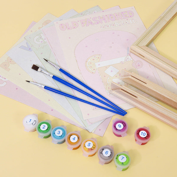 Cocktail Cuties Paint by Numbers Kit by Artspark displaying unpainted cocktail-themed canvases, paint pots, brushes, and wooden frames before painting.
