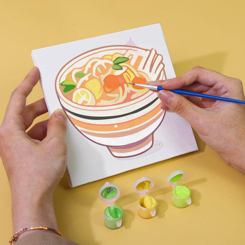 Painting progress of a ramen canvas from the Flavors Around paint by numbers set, with a brush and paint pots displayed.