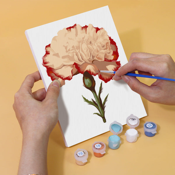 Painting process of a vibrant carnation flower from the Garden Whispers Paint by Numbers Kit. Shows partially painted canvas, paint pots, and brushes from the Artspark kit.